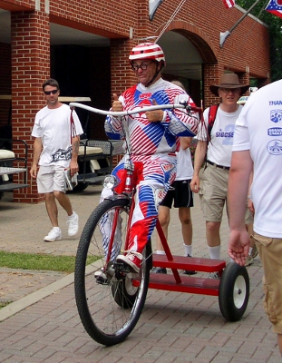 [Man with a striped helmet with a propeller atop it and wearing a red, white, and blue striped costume giving the thumbs up signal while riding atop a super-sized tricycle.]